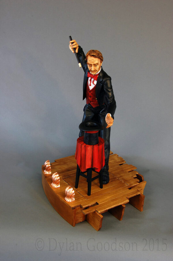 Dylan Goodson's wood carving of a magician standing on stage with wand upraised, about to conjure something out of the hat sitting on the table in front of him.