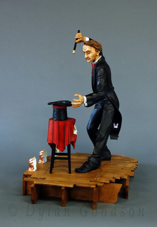 Dylan Goodson's wood carving of a magician standing on stage with wand upraised, about to conjure something out of the hat sitting on the table in front of him.