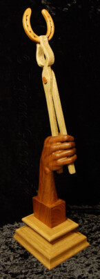 wood sculpture of a hand holding a horseshoe with a pair of blacksmith's tong