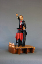Dylan Goodson's wood carving of a magician standing on stage with wand upraised, about to conjure something out of the hat sitting on the table in front of him. Dylan Goodson's wood carving of a magician standing on stage with wand upraised, about to conjure something out of the hat sitting on the table in front of him.