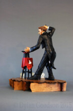 Dylan Goodson's wood carving of a magician standing on stage with wand upraised, about to conjure something out of the hat sitting on the table in front of him.